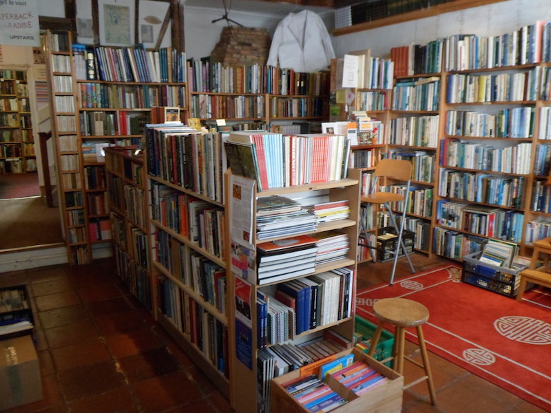 The Topsham Bookshop, ground-floor front room from the door: dense wooden shelves on both sides, parquet floor underfoot, framed images on the back wall, the room receding toward the back-room doorway.